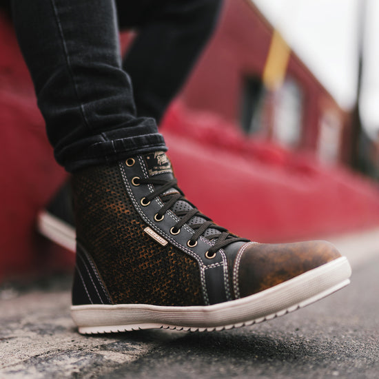 Close-up of a person wearing a brown and black motorcycle sneaker boot on a blurred background