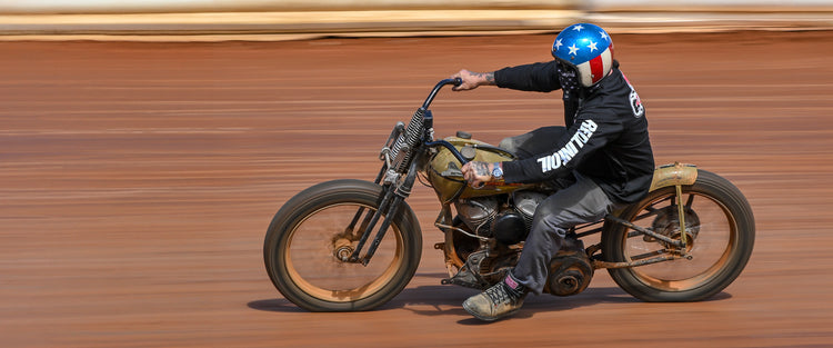 Vintage flat track motorcycle racer competing on a custom board track style motorcycle while wearing classic motorcycle riding apparel, leather motorcycle boots, and a retro helmet inspired by American motorcycle racing culture.