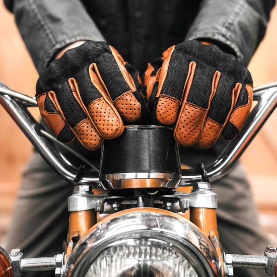 Rider wearing black denim and camel leather motorcycle gloves gripping vintage motorcycle handlebars, showcasing perforated leather fingers and durable riding construction