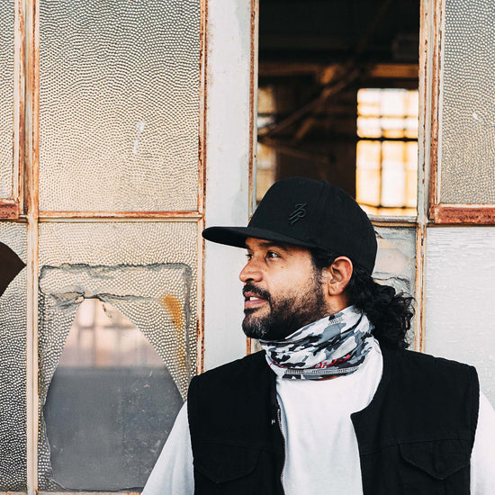Man wearing a black cap and patterned scarf standing in front of a textured wall.