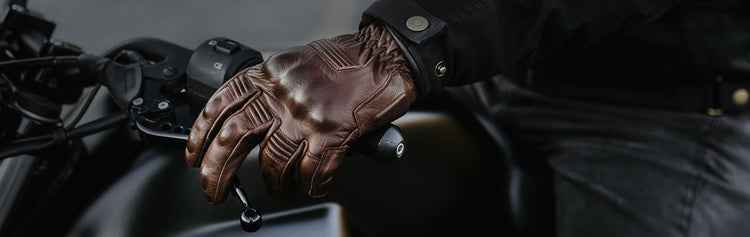 Close-up of a rider wearing a brown leather motorcycle gauntlet glove, resting their hand on the handlebar of a black motorcycle.