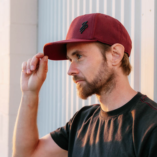 Man wearing a maroon baseball hat with a logo, standing against a light-colored wall.