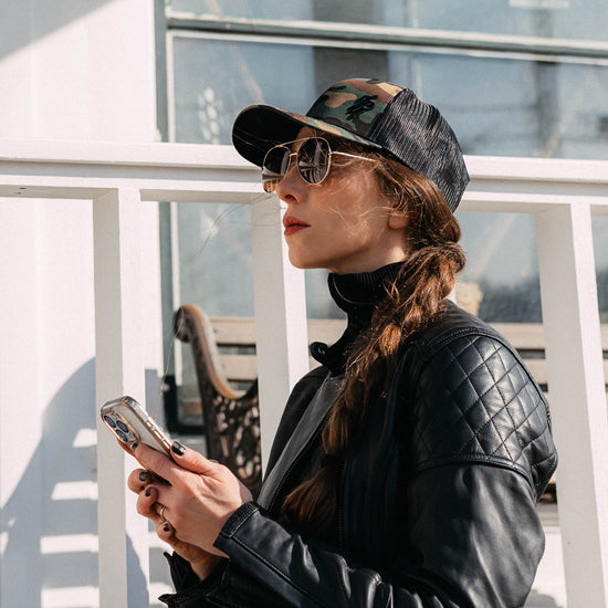 Woman wearing a black quilted jacket, camouflage baseball cap, and sunglasses, looking at her phone.