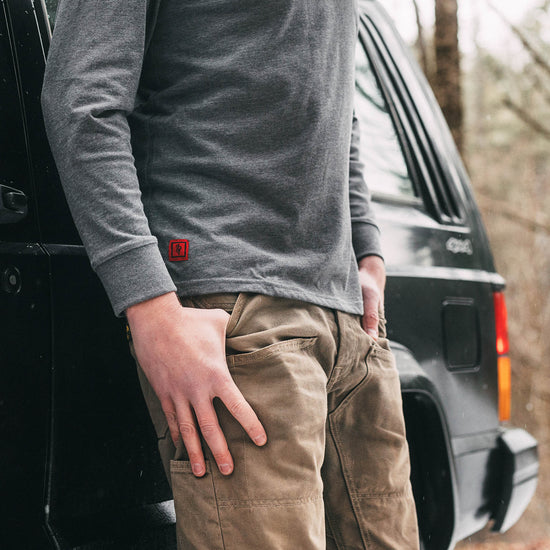 Person wearing a gray long-sleeve shirt with a red logo and brown pants standing next to a black Jeep SUV in a forest setting.