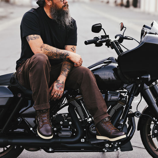 Man sitting on a black Harley-Davidson Road Glide, wearing brown leather motorcycle boots and brown pants, with detailed tattoos visible on his arms and hands.