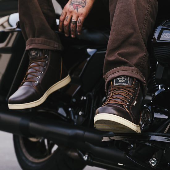 Person wearing brown leather motorcycle boots with visible brand logo on a motorcycle.