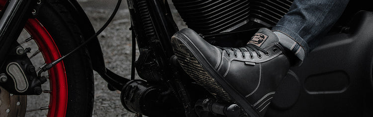 A close-up of a person's foot wearing a black leather motorcycle boot, resting on the footpeg of a black cruiser motorcycle.