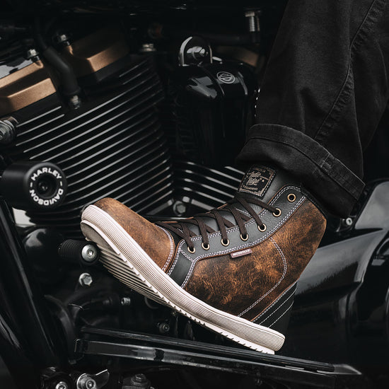 Detailed view of the Indie Ridge Comanche waterproof motorcycle boot against a Harley-Davidson engine, highlighting the brown distressed leather, stitching, and protective design.