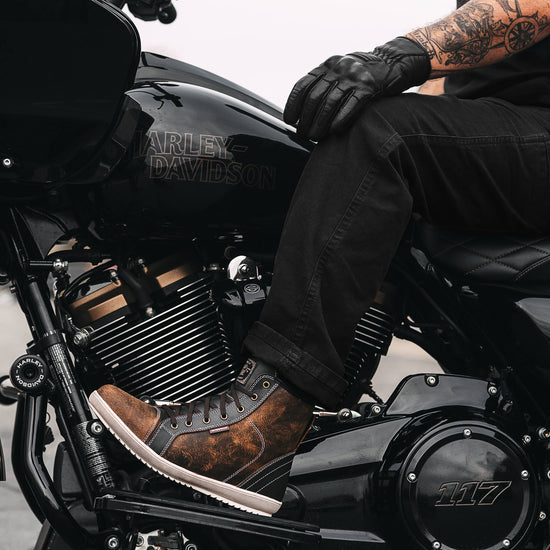 Close-up of the Indie Ridge Comanche waterproof motorcycle boot on a Harley-Davidson rider, showing brown distressed leather and reinforced construction while seated on the bike.