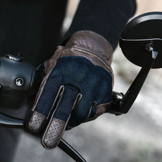Close-up of a brown leather and denim gloved hand on a motorcycle handlebar with a blurred background