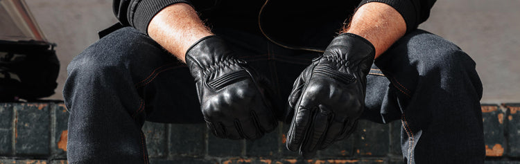 Wide close-up of a seated rider resting their hands between their knees while wearing black leather Indie Ridge Getaway motorcycle gloves.