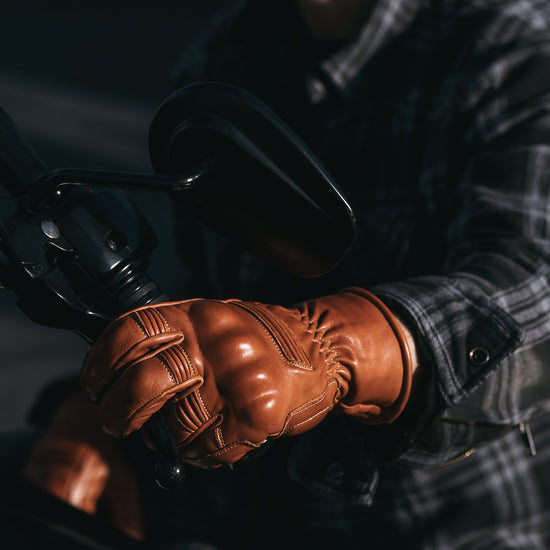 Close-up of a person wearing brown leather gloves on a motorcycle handlebar.