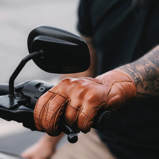 Close-up of a person wearing a golden brown leather glove on a motorcycle handlebar with a blurred background.