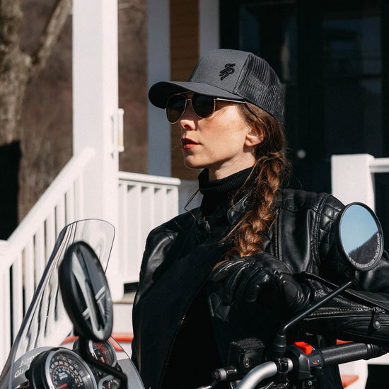 Woman wearing a black leather jacket and gray mesh baseball hat, standing by a motorcycle.