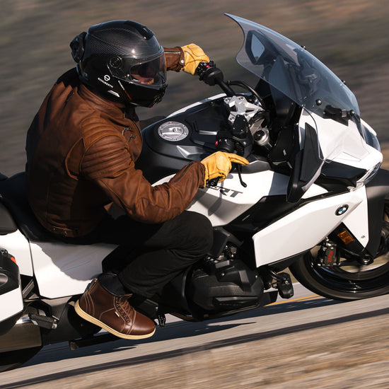 Rider leaning into a turn on a white BMW R1250RS wearing Indie Ridge Apache brown leather boots, tan motorcycle gloves, and a brown leather riding jacket.