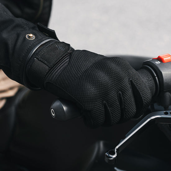 Close-up of Indie Ridge black Powersport motorcycle glove gripping the throttle, reinforced palm and breathable mesh construction visible on the handlebar.