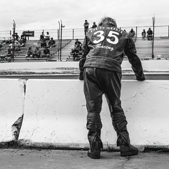 Black and white photo of an Old Soul Racing team member in a leather jacket and Indie Ridge boots looking over the track at New Smyrna Speedway.