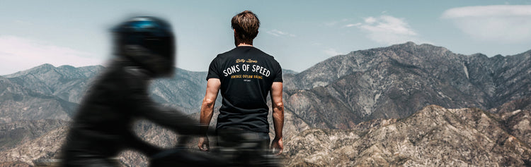 A motorcyclist wearing a Sons of Speed T-shirt overlooks rugged mountain scenery while a blurred motorcycle rides past in the foreground, capturing the speed, freedom, and adventure of motorcycle touring in the American West.
