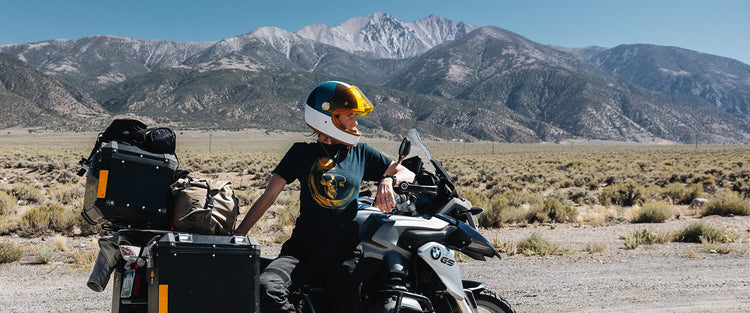 Motorcyclist wearing a navy T-shirt with a yellow skull graphic and brown motorcycle boots beside a loaded BMW GS in the mountains.