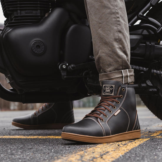 Side view of Indie Ridge warm brown-gray waterproof motorcycle boots, full-grain leather with reinforced soles, shown on a rider beside a Royal Enfield motorcycle.